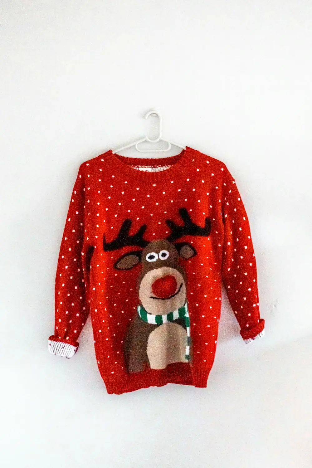 A red knit Christmas sweater with a big reindeer face hanging on a white wall